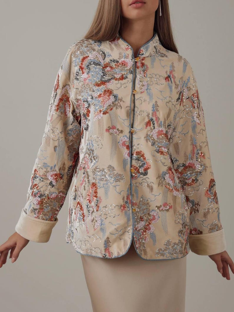 May include: Beige jacket with floral embroidery and sequin details. The jacket features a mandarin collar, long sleeves with rolled cuffs, and gold-colored buttons. The design includes blue trim and a subtle pattern.
