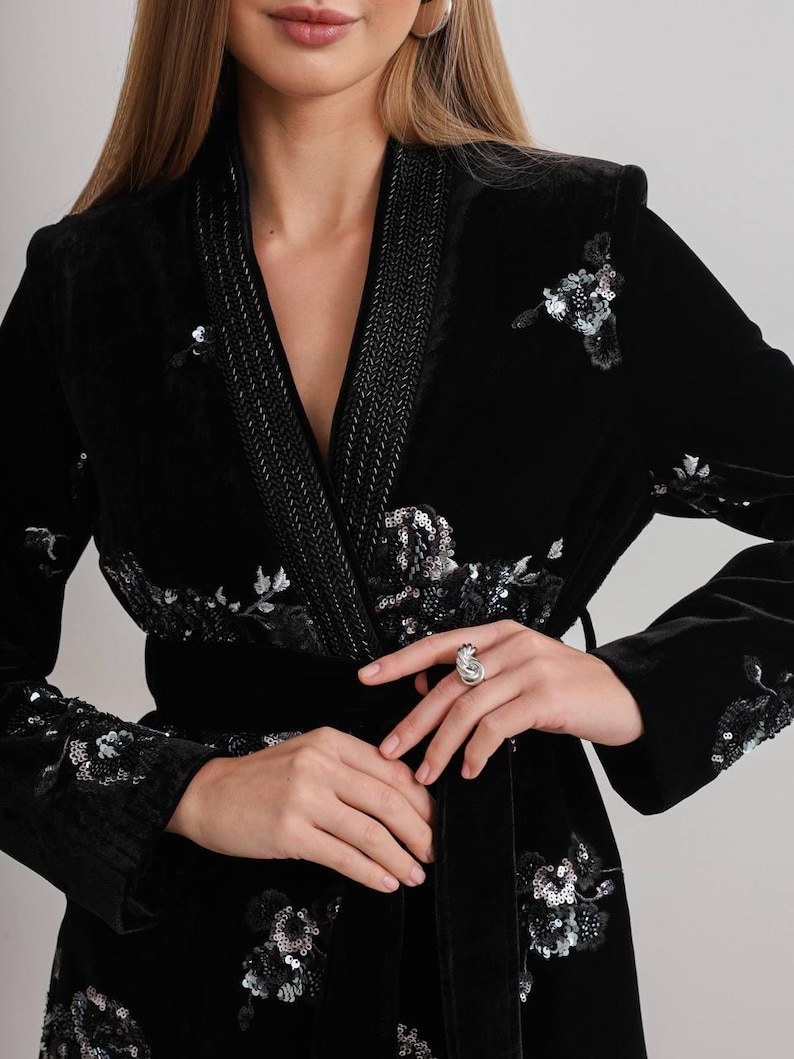May include: Black velvet robe with floral sequin embellishments. The robe features a shawl collar, a braided trim, and a matching belt. The sleeves and front panels are adorned with silver and black sequins in a floral pattern.