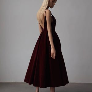 May include: A burgundy-colored formal dress with a V-neckline and a flowing skirt. The dress has thin straps and a low back. The model is wearing silver heels. The dress is knee-length.