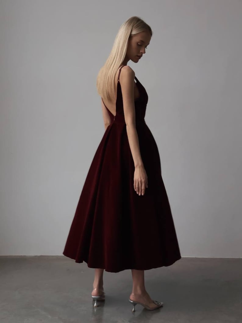 May include: A burgundy-colored formal dress with a V-neckline and a flowing skirt. The dress has thin straps and a low back. The model is wearing silver heels. The dress is knee-length.