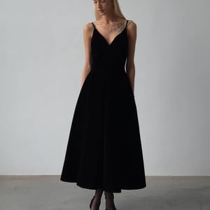 May include: A black midi dress with a V-neckline and spaghetti straps. The dress has a flared skirt and is paired with black heels. The image is set against a plain white background.