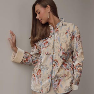 May include: Beige jacket with a mandarin collar and floral embroidery. The jacket features a light blue trim and gold buttons. The sleeves have a cream-colored cuff. The jacket is part of a formal wear collection.