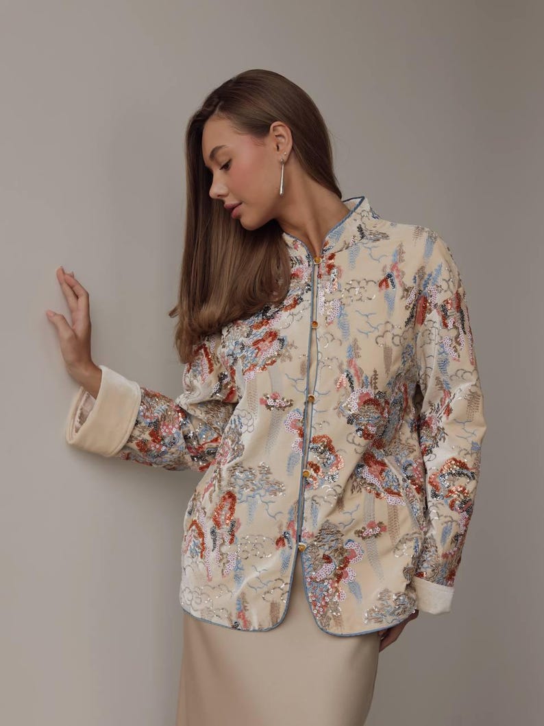 May include: Beige jacket with a mandarin collar and floral embroidery. The jacket features a light blue trim and gold buttons. The sleeves have a cream-colored cuff. The jacket is part of a formal wear collection.
