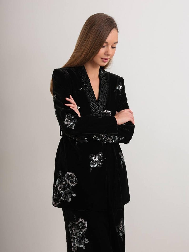 May include: Black velvet jacket and matching skirt set. The jacket features a shawl collar and is embellished with silver floral embroidery. The set is a luxurious, elegant outfit.