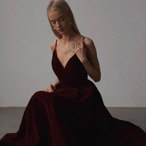 May include: A burgundy velvet gown with a V-neckline and spaghetti straps. The dress has a flowing skirt and is worn by a person sitting on the floor. The dress is a deep, rich color, and the fabric appears soft and luxurious.