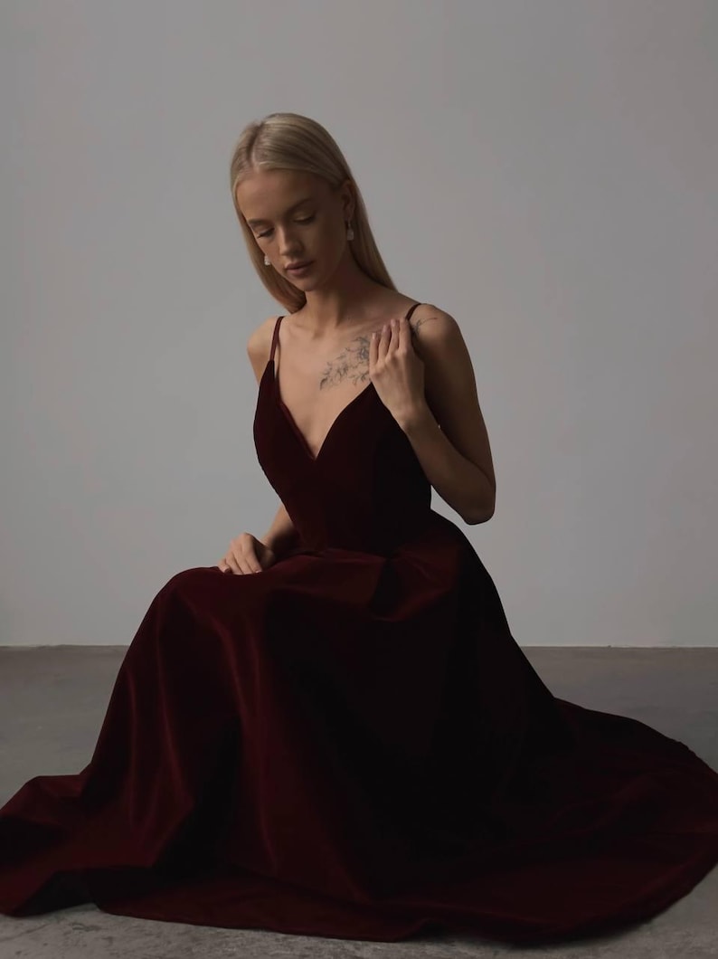May include: A burgundy velvet gown with a V-neckline and spaghetti straps. The dress has a flowing skirt and is worn by a person sitting on the floor. The dress is a deep, rich color, and the fabric appears soft and luxurious.
