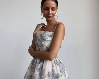 May include: A white floral print dress with a square neckline and spaghetti straps. The dress features a full skirt and a pattern of blue and purple flowers. The model is wearing gold hoop earrings.