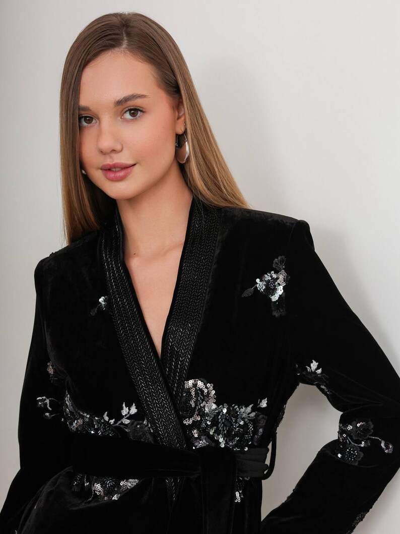May include: A black velvet wrap jacket with floral sequin embellishments. The jacket features a shawl collar, a matching belt, and decorative trim along the lapels. The sleeves are long, and the jacket is a luxurious piece of apparel.