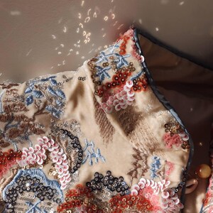 May include: Close-up of a beige jacket collar with intricate embroidery and sequin embellishments. The design features floral and abstract patterns in shades of blue, red, pink, and black. The jacket has a light blue trim.