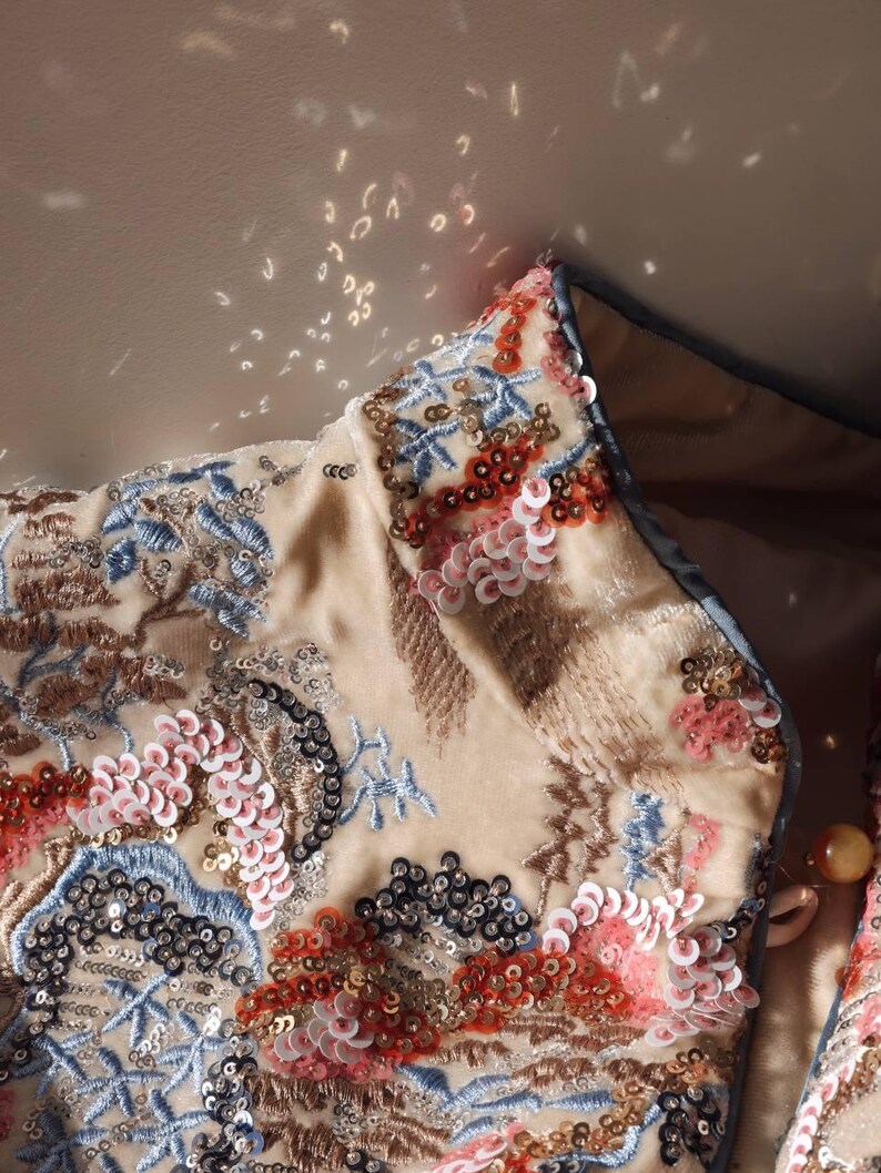 May include: Close-up of a beige jacket collar with intricate embroidery and sequin embellishments. The design features floral and abstract patterns in shades of blue, red, pink, and black. The jacket has a light blue trim.