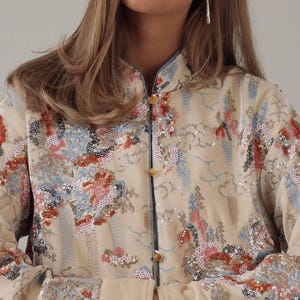May include: Cream-colored jacket with a mandarin collar and floral embroidery. The jacket features a button-up front and is embellished with sequins in shades of red, blue, and silver. The sleeves have a light blue trim.