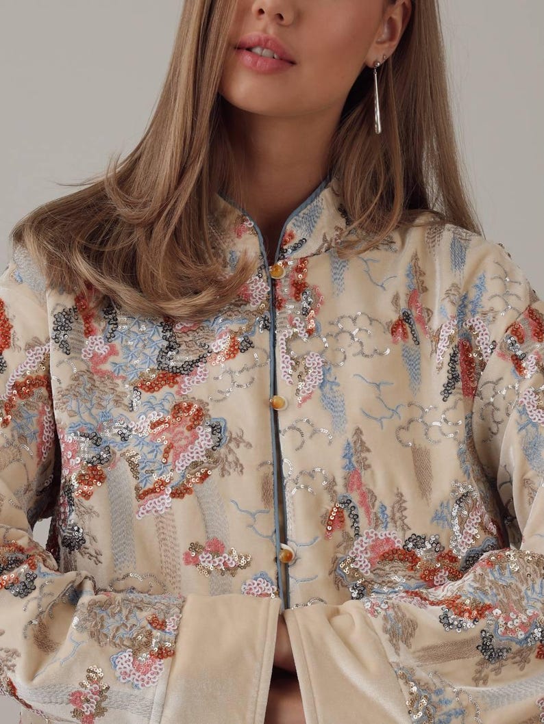 May include: Cream-colored jacket with a mandarin collar and floral embroidery. The jacket features a button-up front and is embellished with sequins in shades of red, blue, and silver. The sleeves have a light blue trim.