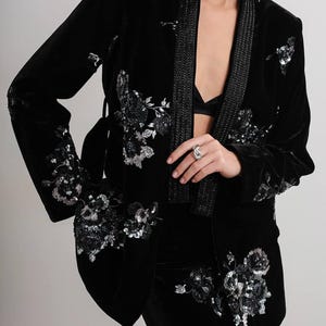 May include: Black velvet blazer with floral sequin embellishments. The blazer features a shawl collar with a beaded trim and a matching belt. The sleeves and front of the blazer are adorned with silver sequin floral designs. The blazer is part of a matching set.