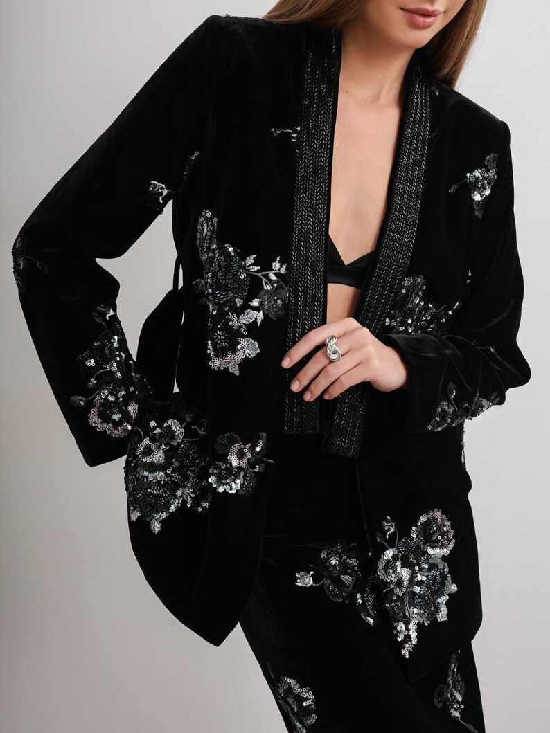 May include: Black velvet blazer with floral sequin embellishments. The blazer features a shawl collar with a beaded trim and a matching belt. The sleeves and front of the blazer are adorned with silver sequin floral designs. The blazer is part of a matching set.