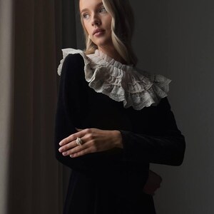 May include: A woman wearing a black dress with a white lace collar and a ring. The dress has long sleeves and a high neckline. The collar is layered and ruffled. The ring is a cluster of small stones.