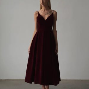 May include: A burgundy midi dress with a V-neckline and spaghetti straps. The dress has a fitted bodice and a flared skirt. The fabric appears to be velvet or a similar material. The dress is suitable for formal occasions.