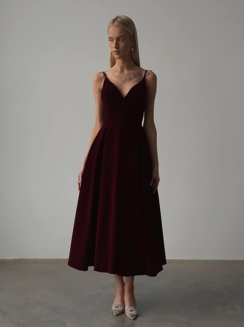 May include: A burgundy midi dress with a V-neckline and spaghetti straps. The dress has a fitted bodice and a flared skirt. The fabric appears to be velvet or a similar material. The dress is suitable for formal occasions.