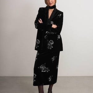 May include: A black velvet suit with floral embroidery. The suit includes a long jacket and a knee-length skirt. The jacket has a shawl collar and the skirt has a straight silhouette. The suit is worn with black shoes.