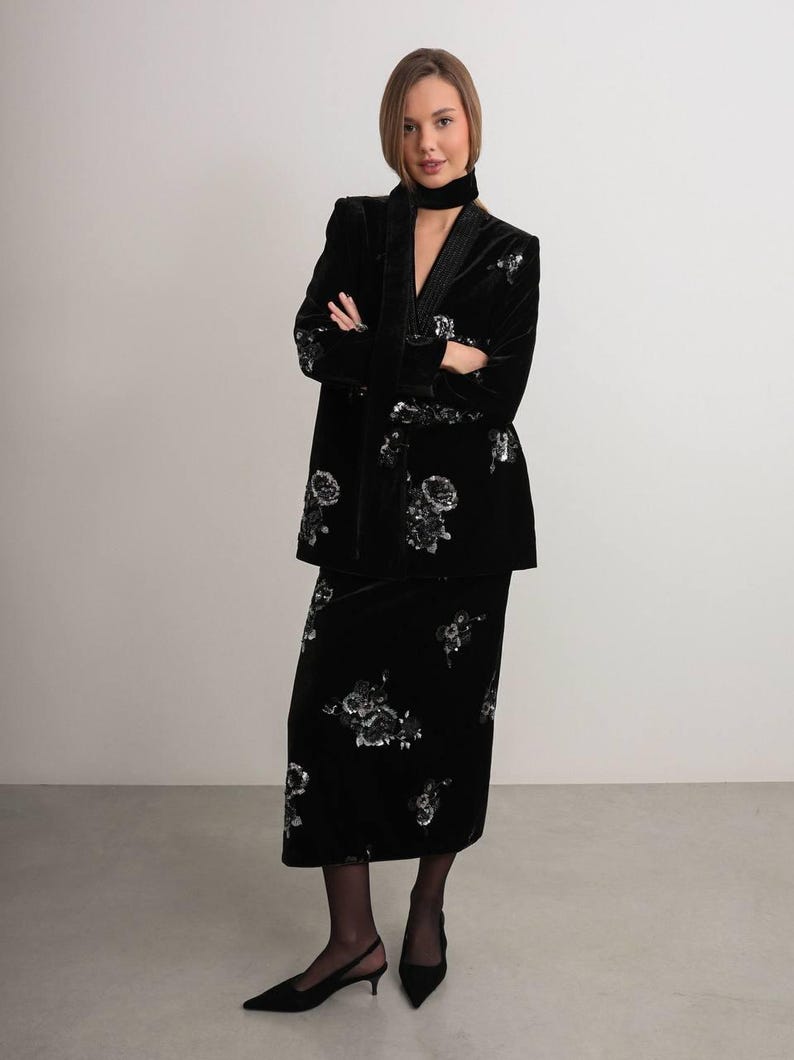 May include: A black velvet suit with floral embroidery. The suit includes a long jacket and a knee-length skirt. The jacket has a shawl collar and the skirt has a straight silhouette. The suit is worn with black shoes.