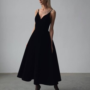 May include: A black midi dress with a V-neckline and spaghetti straps. The dress has a flared skirt and is paired with black heels. The dress is made of a soft, flowing fabric, perfect for a formal event or special occasion.