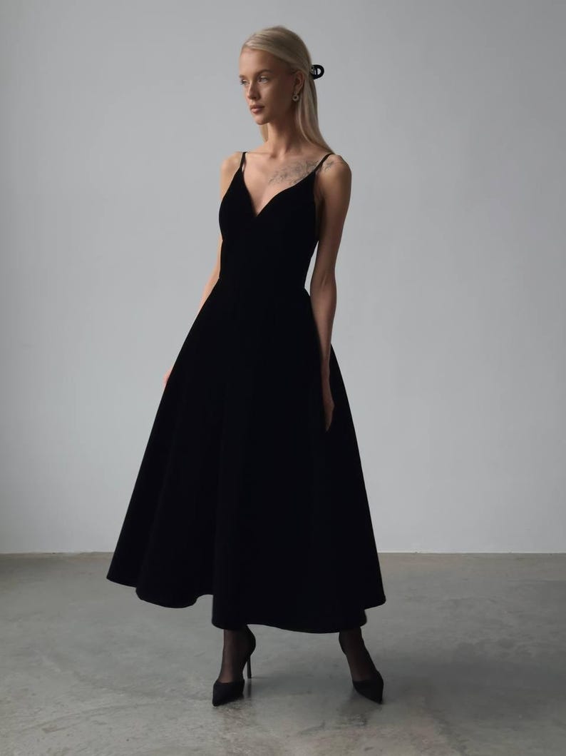 May include: A black midi dress with a V-neckline and spaghetti straps. The dress has a flared skirt and is paired with black heels. The dress is made of a soft, flowing fabric, perfect for a formal event or special occasion.