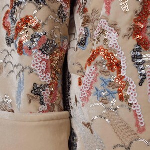 May include: Close-up of a cream-colored jacket with intricate embroidery and sequin embellishments. The design features floral and landscape motifs in shades of red, pink, blue, and silver. The jacket has a classic tailored cut.