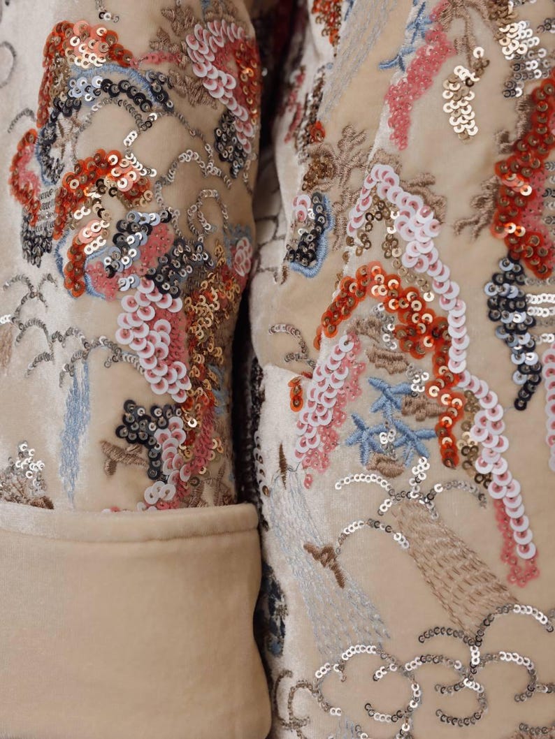 May include: Close-up of a cream-colored jacket with intricate embroidery and sequin embellishments. The design features floral and landscape motifs in shades of red, pink, blue, and silver. The jacket has a classic tailored cut.
