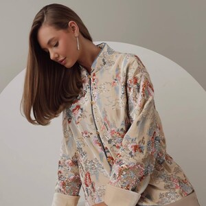 May include: A beige, long-sleeved jacket with a floral pattern in shades of red, blue, and pink. The jacket has a mandarin collar and button closures. The cuffs and collar are trimmed with a lighter beige fabric. The jacket is part of a clothing collection.
