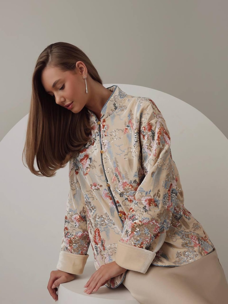 May include: A beige, long-sleeved jacket with a floral pattern in shades of red, blue, and pink. The jacket has a mandarin collar and button closures. The cuffs and collar are trimmed with a lighter beige fabric. The jacket is part of a clothing collection.
