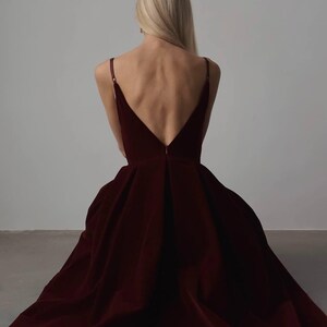 May include: A burgundy velvet gown with a deep V-neckline and spaghetti straps. The dress features a full, flowing skirt that drapes to the floor. The back of the dress is open, showcasing the wearer's back. The model has long blonde hair.