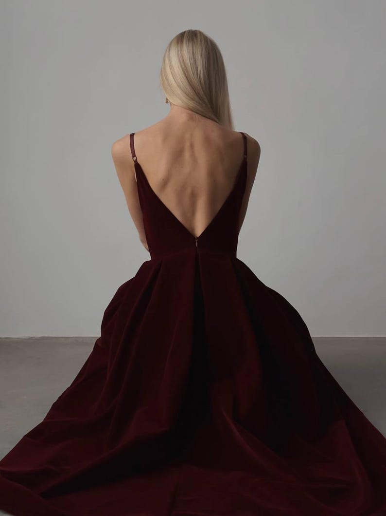 May include: A burgundy velvet gown with a deep V-neckline and spaghetti straps. The dress features a full, flowing skirt that drapes to the floor. The back of the dress is open, showcasing the wearer's back. The model has long blonde hair.