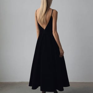 May include: A black, floor-length dress with a V-neckline and spaghetti straps. The dress has a flared skirt and is worn with black heels. A black hair clip is visible in the model's blonde hair.