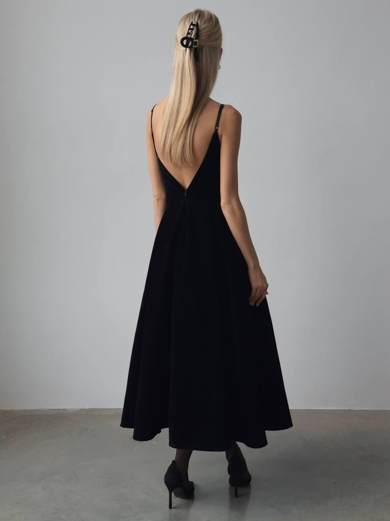 May include: A black, floor-length dress with a V-neckline and spaghetti straps. The dress has a flared skirt and is worn with black heels. A black hair clip is visible in the model's blonde hair.