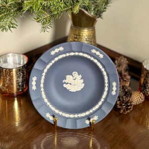 May include: A blue decorative plate with a white relief design of a classical scene. The plate has a scalloped edge and is displayed on a gold stand. The plate is surrounded by pine cones, candles, and a vase with evergreen branches.