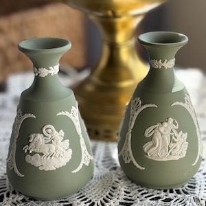 May include: Two sage green decorative vases with white relief designs. The vases have a classic shape with a flared top and a detailed relief of figures and a chariot. The vases are displayed on a white lace doily.