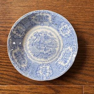 May include: A round ceramic saucer featuring a blue and white transferware pattern. The center depicts a landscape with buildings, trees, and mountains, while the scalloped rim is decorated with floral motifs and additional architectural scenes.