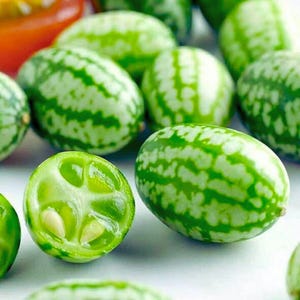 May include: A collection of small, green, watermelon-like fruits with light green and white striped exteriors. One fruit is cut open, revealing a translucent interior and small seeds. The fruits are clustered together, with a few red tomatoes in the background.