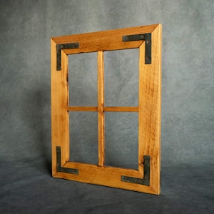 Puede incluir: Un marco de ventana de madera rústica con un acabado natural. El marco presenta un patrón de cuadrícula y está reforzado con escuadras de metal oscuro. La veta de la madera es visible y el marco está ligeramente inclinado.