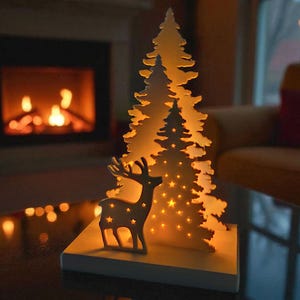 May include: A decorative Christmas tree and reindeer silhouette illuminated by warm lights. The tree and reindeer are made of a light-colored material and sit on a white base. The background features a fireplace and soft lighting.