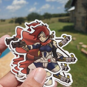 Può includere: Una pila di adesivi raffiguranti un personaggio guerriero femminile con capelli rossi, un mantello rosso e un'armatura futuristica. Il personaggio tiene in mano due pistole e ha diverse armi attaccate alle braccia. Gli adesivi sono tenuti in mano su uno sfondo di cielo blu ed erba verde.