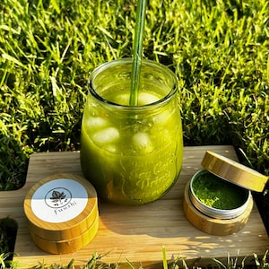 May include: A refreshing green beverage in a glass jar with ice cubes and a straw, alongside a small container of green powder and a wooden container with the text "Fuushi". The items are on a wooden board, set against a backdrop of green grass.