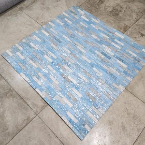 Puede incluir: Una alfombra cuadrada con un patrón de ladrillos azules, blancos y grises. La alfombra está hecha de un material duradero y es adecuada para uso en interiores o exteriores. La alfombra está colocada sobre un suelo de baldosas grises.