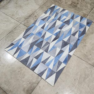 Könnte beinhalten: Rechteckiger Teppich mit geometrischem Muster in Blau, Grau und Weiß. Das Design besteht aus sich wiederholenden Dreiecken. Der Teppich ist aus weichem Material und für ein modernes Interieur geeignet. Maße ca. 150 cm x 200 cm.