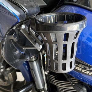 May include: A black motorcycle cup holder mounted on a blue motorcycle. The cup holder is made of a dark gray plastic with a cage-like design. The mounting hardware is visible, with bolts and a pivoting arm.