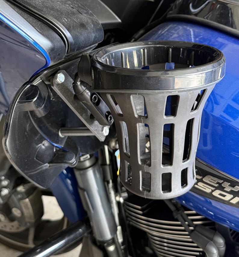 Puede incluir: Un portavasos negro para motocicleta montado en una motocicleta azul. El portavasos es de pl&aacute;stico gris oscuro con un dise&ntilde;o similar a una jaula. El hardware de montaje es visible, con pernos y un brazo giratorio.