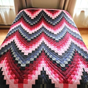 May include: A handmade quilt with a geometric chevron pattern in shades of pink, red, black, and gray. The quilt is spread across a bed, showcasing its intricate design and craftsmanship. The quilt's colors create a visually striking and cozy aesthetic.