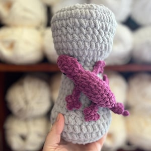 May include: A handmade crochet lizard in shades of gray and purple. The lizard is wrapped around a gray, cylindrical shape. The item is made of soft yarn and has a textured appearance. The background features shelves with white yarn.