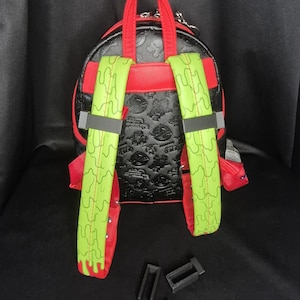 May include: Black and red mini backpack with green straps. The backpack has a repeating pattern on the front and sides. The straps are green with a wavy design. The top handle and trim are red.