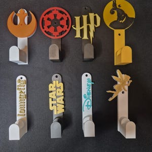 May include: A selection of decorative wall hooks with diverse designs. The hooks feature Star Wars, Harry Potter, and Disney themes, alongside other unique shapes and logos. Each hook has a coloured design attached to a white or grey hook base.