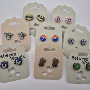 Può includere: Una collezione di sette paia di orecchini a lobo presentati su cartoncini beige individuali, ognuno con disegni di personaggi della cultura pop come Batman, Captain America, Grogu, Wonder Woman, Mandalorian, Sonic, Groot e le Tartarughe Ninja.